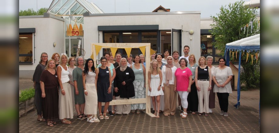 50 Jahre Kita Neuerburg Das Foto zeigt Mitarbeiter der Kindertagesstätte vor dem Eingang des Gebäudes. Sie halten einen gebastelten Rahmen in den Händen und eine gebastelte Krone ist zu sehen.