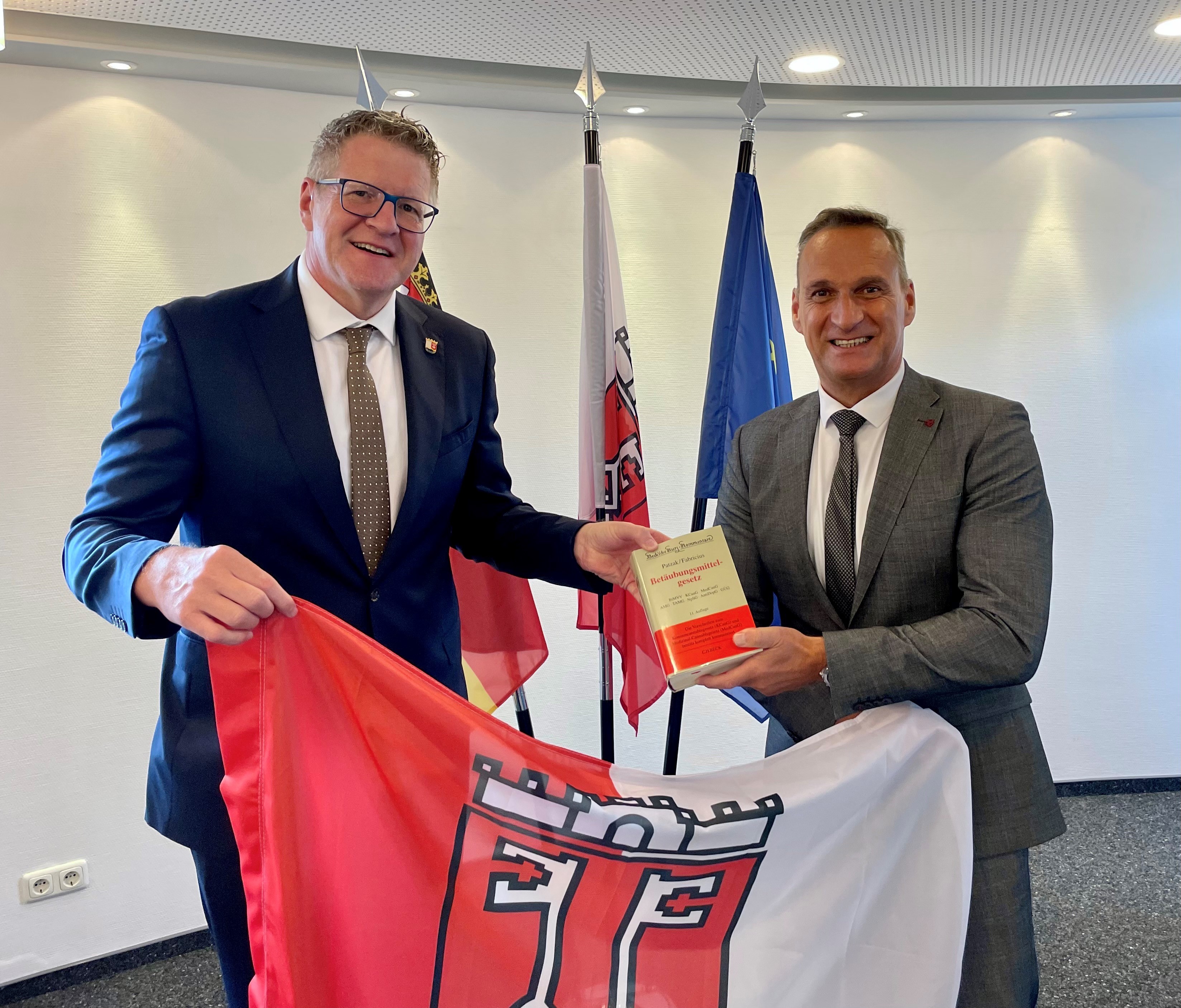 Das Foto zeigt Jörn Patzak, den Leiter der JVa Wittlich und Bürgermeister Rodenkirch bei der Übergabe der Fahne der Stadt Wittlich.