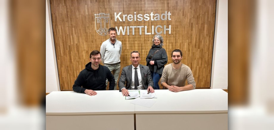 Bürgermeister Joachim Rodenkirch und die beide Brüder Niklas und Jonas Butzen unterzeichnen den Pachtvertrag für den Wohnmobilstellplatz Zweibächen in Wittlich. Bei der Unterzeichnung waren der Fachbereichsleiter Rainer Wener (hinten stehend links) und die Leiterin der Tourist-Information Wittlich Nicole Baller mit dabei (Foto: Sebastian Klein.)