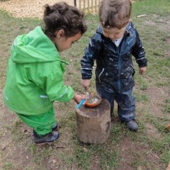 Das Bild zeigt zwei Kinder auf dem Außengelände mit Matschkleidung die vor einem kurzen Baumstumpf stehen. Darauf steht eine kleine Schüssel mit Matsch. Die Kinder haben jeweils einen Pinsel in der Hand und tauchen ihn in den Matsch, dabei schauen sie nach unten zur Schüssel.