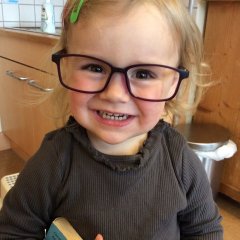 Das Bild zeigt ein kleines Mädchen das eine große lila Brille trägt. Es lächelt in die Kamera.