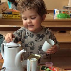 Das Bild zeigt einen kleinen Jungen der eine Tasse in der Hand hält. Mit der anderen Hand hebt er den Deckel einer Teekanne an die auf dem Tisch vor ihm steht.