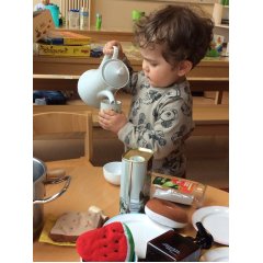 Auf dem Bild ist ein kleiner Junge zu sehen der sehr konzentriert eine Teekanne in der einen und eine Teetasse in der anderen Hand festhält. Er spielt, dass er etwas aus der Teekanne in die Teetasse umfüllt. Vor ihm steht ein kleiner Tisch auf dem Töpfe, Teller und Essen aus Stoff liegen.