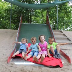 Das Bild zeigt vier Kinder die auf einer roten Decke am Ende der Rutsche nebeneinander sitzen. Ein Mädchen streckt dabei die Zunge raus.