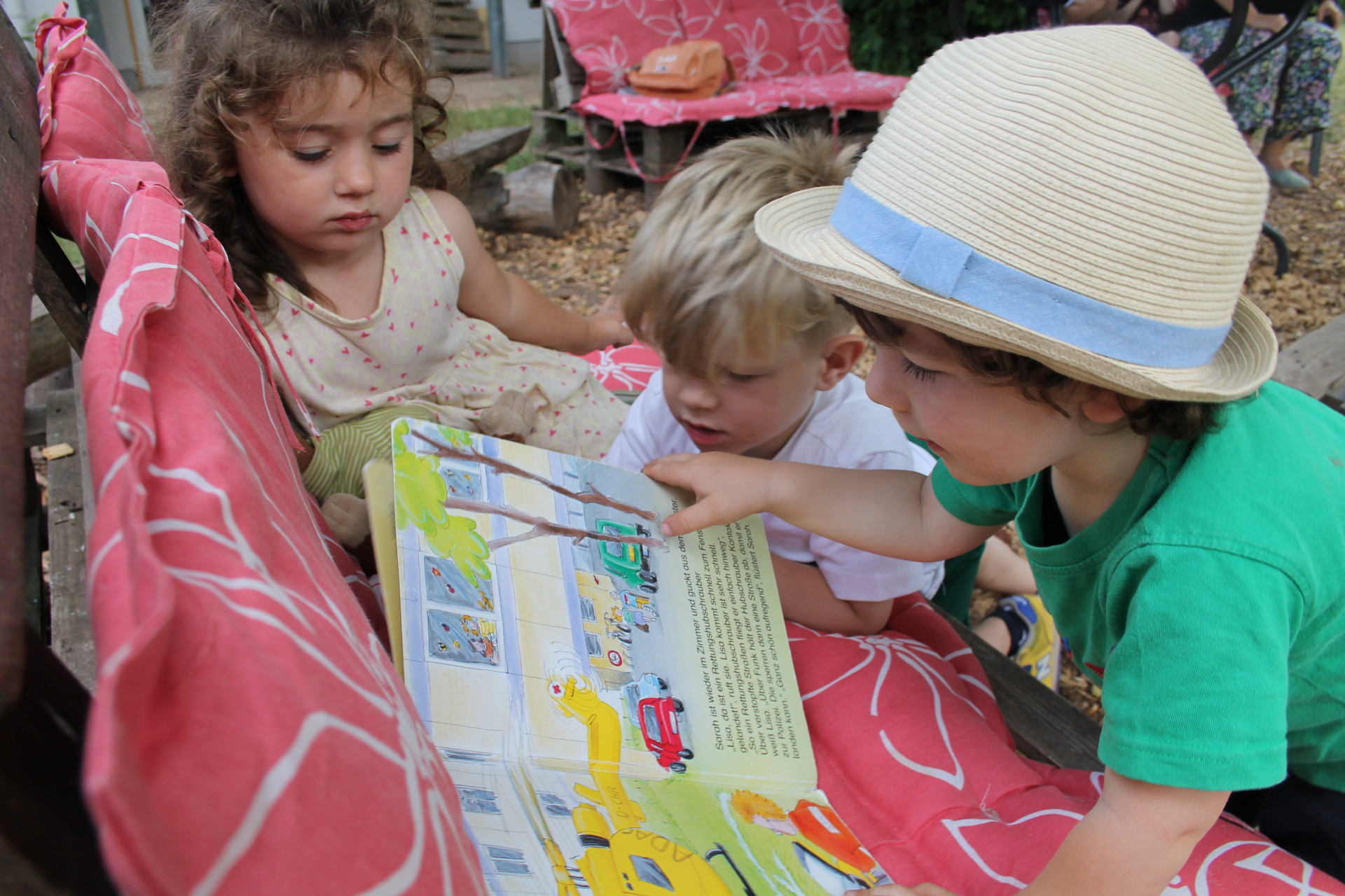Buch Forscher Dieses Bild zeigt drei Kinder, die sich ein Buch über den Rettungsdienst anschauen. Dabei sitzen sie auf einer Bank im Außengelände.
