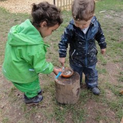 Das Bild zeigt einen kleinen Jungen und ein Mädchen mit Matschkleidung die nebeneinander stehen. Sie sind über einen Baumstamm gebeugt, auf dem eine kleine Schüssel mit Matsch steht. Die Kinder haben einen Pinsel in der Hand den sie in den Matsch tauchen. 