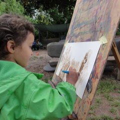 Das Bild zeigt ein kleines Mädchen mit Matschkleidung, dass vor einer Staffelei draußen steht. Sie malt mit einem Pinsel Matsch auf ein weißes Papier an der Staffelei. 