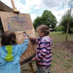 Das Bild zeigt zwei Mädchen mit Matschkleidung die vor einer Staffelei auf dem Außengelände stehen. Sie malen mit Pinseln braunen Matsch auf ein weißes Blatt Papier an der Staffelei.