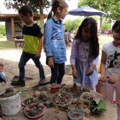 Das Bild zeigt sechs Kinder die um einen kleinen Tisch im Sandkasten stehen. Auf dem Tisch stehen Töpfe und Schüsseln gefüllt mit Sand, Blättern und Blumen.