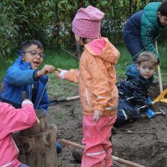 Das Bild zeigt fünf Kinder in Matschkleidung auf dem Matschhügel. Ein Mädchen im Vordergrund hämmert einen Nagel in einen Baumstamm. Hinter ihr gibt ein Junge einem Mädchen einen Nagel in die Hand. Zwei Jungs sind im Hintergrund mit einem Hammer und einer Schaufel am arbeiten.