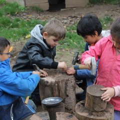 Das Foto zeigt vier Kinder um einen Baumstamm. Die Kinder haben einen Hammer und Nägel in der Hand und hämmern diese in den Baumstamm. 