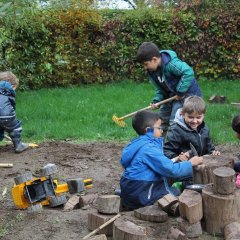 Das Bild zeigt sechs Kinder in Matschkleidung auf dem Matschhügel. Zwei der Kinder graben mit Schaufel und Rechen . Vier Kinder sitzen um einen Baumstamm und nageln Nägel hinein.