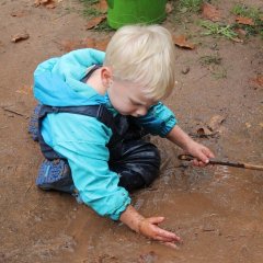 Das Bild zeigt einen Jungen in Matschkleidung der in einer Pfütze kniet. In der einen Hand hält er einen Stock. Seine andere Hand und den Arm hält er in die Pfütze und staut das Wasser.  Er schaut dabei nach unten auf seine Hand.