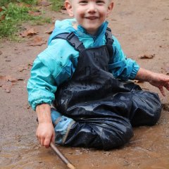 Das Bild zeigt einen Jungen mit Matschkleidung. Er sitzt auf dem Boden in einer Matschpfütze und lächelt in die Kamera. Im Gesicht hat er viele Matschspritzer verteilt. In der einen Hand hält er einen Stock.