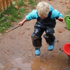 Auf dem Bild sieht man einen Jungen in Matschkleidung der in einer Matschpfütze hoch springt. Er schaut dabei zum Boden. In der Hand hält er einen Stock.