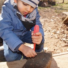 Ein Junge hält einen roten Schraubenzieher in der linken Hand und eine Schraube in der rechten Hand und versucht diese in ein Stück Holz zu schrauben