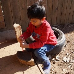 Ein Junge sitzt auf einem Reifen und hat ein Stück Holz vor sich. Er dreht mit einem roten Schraubenzieher eine Schraube in das Stück Holz. 