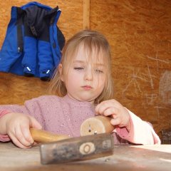 Ein Mädchen sitzt vor einem Stück Holz und hält mit der einen Hand einen Nagel fest. Mit der anderen Hand hält sie den Hammer.