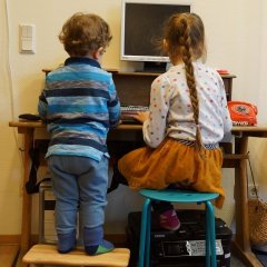 Ein kleiner Junge steht auf einem Hocker und ein großes Mädchen sitzt auf einem Stuhl am Schreibtisch. Beide schauen auf den PC-Bildschirm der vor ihnen steht. Das Mädchen tippt mit den Fingern auf der Tastatur.