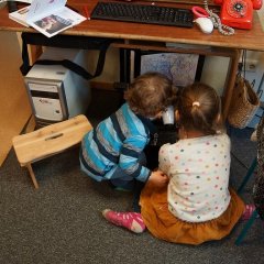 Ein kleiner Junge und ein großes Mädchen sitzen zusammen vor einem Drucker auf dem Boden. Der Junge legt den Föhn auf den Scanner des Druckers.