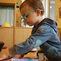 Auf dem Bild sieht man einen Jungen mit einer großen grünen Brille der an einem Tisch steht und ein Messer in der einen Hand hat, mit der anderen greift er nach etwas. Vor ihm auf dem Tisch liegt ein Schreibheft und verschiedenes Essen aus Stoff und Holz. Der Junge schaut konzentriert au den Tisch.