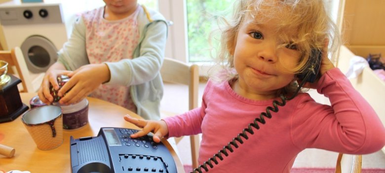 Auf dem Bild sieht man ein kleines Mädchen, dass ein Kabeltelefon vor sich hat. Den Hörer hält sie mit der linken Hand am Ohr und schaut in die Kamera. Mit der rechten Hand tippt sie auf den Tasten des Telefons. Im Hintergrund öffnet ein Mädchen eine Dose in Form eines Muffins.