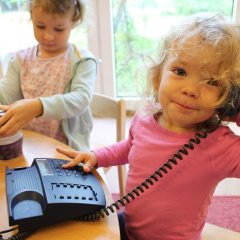 Auf dem Bild sieht man ein kleines Mädchen, dass ein Kabeltelefon vor sich hat. Den Hörer hält sie mit der linken Hand am Ohr und schaut in die Kamera. Mit der rechten Hand tippt sie auf den Tasten des Telefons. Im Hintergrund öffnet ein Mädchen eine Dose in Form eines Muffins.