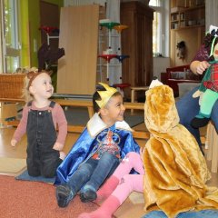 Auf dem Bild sieht man eine Erzieherin mit einer Handpuppe in der Hand. Vor ihr sitzen Kinder im Kreis mit  Kostümen als Prinz, Rentier, Igel und Tiger. Sie schauen lachend zur Erzieherin. 
