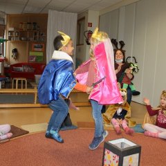 Auf dem Bild sieht man zwei Kinder ein Junge mit einem Prinzenkostüm und ein Mädchen mit einem Prinzessinenkostüm. Die Kinder halten sich an den Händen und hüpfen in die Luft. Hinter ihnen sitzen Kinder im Kreis und eine Erzieherin, sie singen und klatschen.