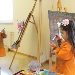 Das Bild zeigt ein Mädchen vor einem großen Spiegel. Rechts neben ihm steht eine Staffelei und links ein kleiner Tisch mit Wasserfarben. Das Mädchen hält einen Pinsel in der rechten Hand und malt sich damit die linke Hand an. Es schaut dabei nach unten auf beide Hände.