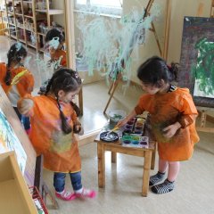 Auf dem Bild sind zwei Mädchen im Malraum zu sehen. Sie tragen beide Malkittel und stehen vor einem großen Spiegel der mit Farbe verschmiert ist. Rechts und links neben den Kindern befindet sich jeweils eine Staffelei mit einem Blatt Papier drauf, dort haben die Kinder schon mit bunter Farbe gemalt. Das linke Kind schaut auf den Farbkasten der auf dem Tisch zwischen en Kinder steht und malt mit der rechten Hand auf das Blatt Papier. Das rechte Kind steht vor dem Tisch und tunkt ihren Pinsel in die Farben.