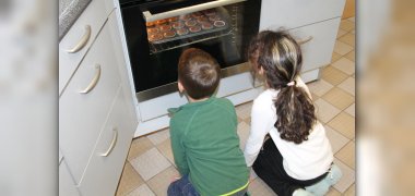 Auf dem Bild sitzen zwei Kinder vor dem Backofen in der Küche und schauen auf die Muffins im Backofen.