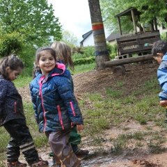 Vier Kinder mit Matschkleidung stehen in einer Matschpfütze. Die Kinder sind von unten bis oben mit Matsch verschmiert und lachen.