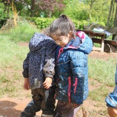 Zwei Kinder stehen in einer Matschpfütze mit Matschkleidung. Beide sind von unten bis oben mit Matsch verschmiert. Sie laufen aneinander vorbei und schauen nach unten.