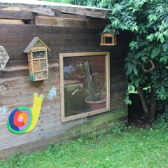 Auf dem Bild ist die Werkstatt zu sehen. Durch eine Plexiglasscheibe kann man in das Innere herein- oder herausschauen. Man sieht einen Teil der Werkbank. An der Außenwand hängt eine große, bunte Schnecke aus Holz. Darüber befinden sich zwei Insektenhotels, einmal als Form eines Häuschen auf dem in bunter Schrift das Wort Insektenhotel steht und einmal als sechseckige Form. Rechts befindet sich ein grüner Busch an dem ein Vogelhaus und ein geflochtener Weidenkranz hängt. 