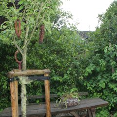 Auf dem Bild ist im Vordergrund ein Baum zu sehen, an dem geflochtene Weidenkränze hängen. Im Hintergrund steht ein Holztisch auf dem ein Gesteck steht.