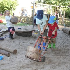 Auf dem Bild sieht man wie zwei Kinder mit einer Sackkarre zwei dicke Holzscheiben durch den Sand schieben. Ein Mädchen hebt eine dicke Holzscheibe mit den Händen hoch und geht neben ihnen durch den Sand.