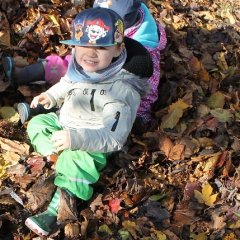 Ein Kind sitzt mit seiner Matschkleidung und Gummistiefeln in einem Laubhaufen und grinst in die Kamera.