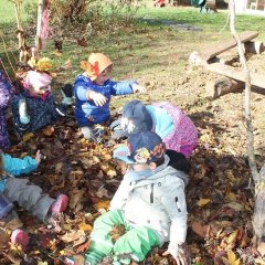 Auf dem Bild sieht man sechs Kinder im Laub sitzen mit Matschkleidung und Gummistiefeln. Sie werfen das Laub in die Luft über sich und lachen dabei. 
