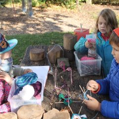 Auf dem Bild sieht man vier Kinder an einem Holztisch sitzen. Auf dem Tisch liegen Holzscheiben und durchsichtige Kisten mit Wolle. In der Mitte der Kinder liegen Figuren aus Ästen, Kastanien und Wolle. Ein Junge vorne rechts wickelt blaue Wolle und eine Figur aus Kastanie und Zahnstocher. 