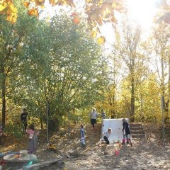 Auf dem Bild sieht man eine Rutsche mit einer Treppe hinauf, Kinder rutschen hinunter und laufen den Hügel daneben hoch und runter. Im Vordergrund steht ein Tisch im Sandkasten mit Bänken daran. Ein paar Kinder laufen auf dem Hügel zwischen den Bäumen mit bunten Blättern hindurch.