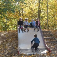 Auf dem Bild sieht man vier Kinder nebeneinander auf der Rutsche sitzen. Ein Kind kniet auf allen vieren unten am Ende der Rutsche.