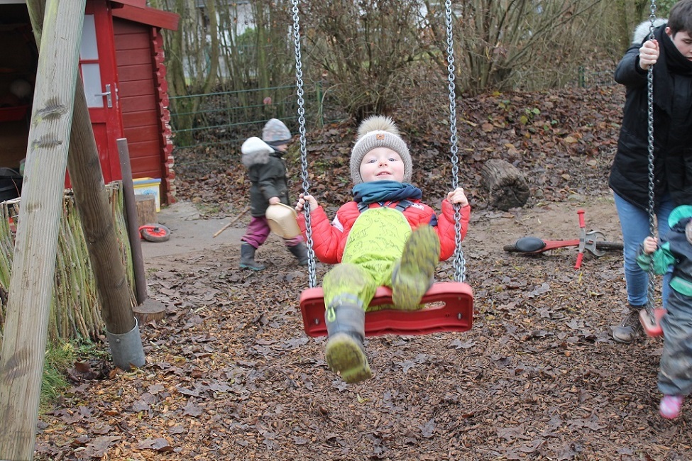 Auf dem Bild ist ein kleiner Junge mit Matschkleidung auf der Schaukel am schaukeln. Er lächelt in die Kamera. Neben ihm steht ein Erzieher hinter der Schaukel und vor der Schaukel steht ein Kind.