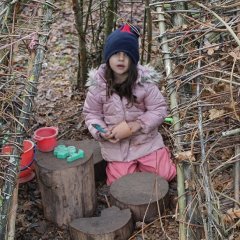 Auf dem Bild ist ein Mädchen im Weidentipi in Matschkleidung zu sehen, dass hinter drei unterschiedlich großen Baumstämmen sitzt. Sie hat einen Becher und ein Plastikmesser in der Hand. Auf dem Baumstamm liegt ein kleiner roter Becher und ein Eisenbahn-Förmchen. Daneben steht ein roter großer Eimer.