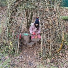 Auf dem Bild ist ein Weidentipi zu sehen. In dem Tipi sitzt ein Mädchen mit Matschkleidung. Vor ihm liegen drei unterschiedlich hohe Baumstämme. Auf den Baumstämmen steht ein Becher und ein Förmchen. Das Mädchen schüttet mit einem Becher in der Hand etwas in den Becher auf dem Baumstamm. 