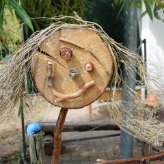 Auf dem Bild ist ein Holzmensch zu sehen. Das Gesicht besteht aus einer Holzscheibe, die Augen aus Kastanien, die Nase aus einem kleinen Tannenzapfen, der Mund und die Augenbrauen aus gebogenen Stöcken und die Haare aus dünnen Ästen. Der Kopf steht auf einem dünnen Stock als Hals. 