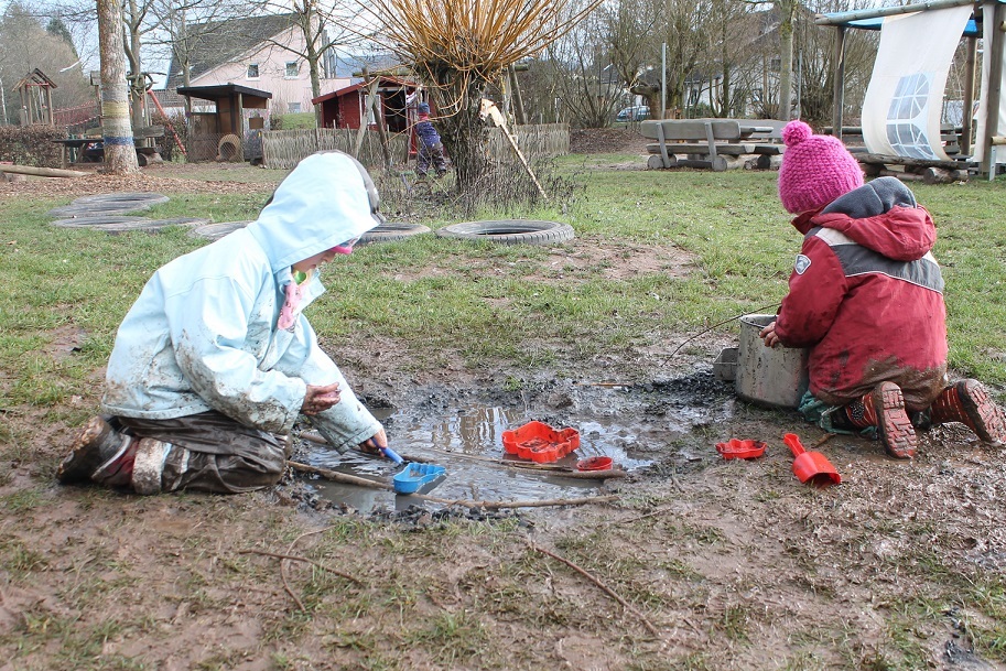 Zwei Kinder knien mit Matschkleidung vor einer Matschpfütze. Das eine Kind füllt mit einem Löffel Matsch in ein Förmchen. Das andere Kind rührt in einem großen Topf.