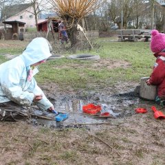 Zwei Kinder knien mit Matschkleidung vor einer Matschpfütze. Das eine Kind füllt mit einem Löffel Matsch in ein Förmchen. Das andere Kind rührt in einem großen Topf.