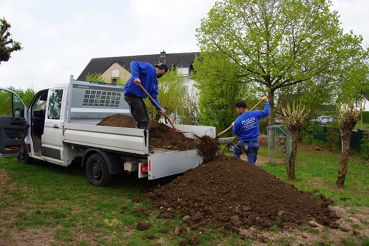 Zwei Arbeiter der Firma Meter stehen auf der Ladefläche des Autos und der Wiese und schaufeln den Boden auf die Wiese.