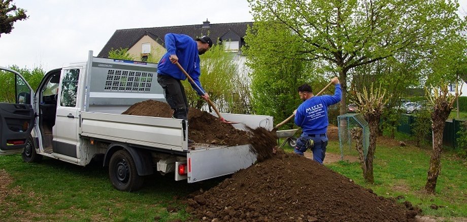 Zwei Arbeiter der Firma Meter stehen auf der Ladefläche des Autos und der Wiese und schaufeln den Boden auf die Wiese.